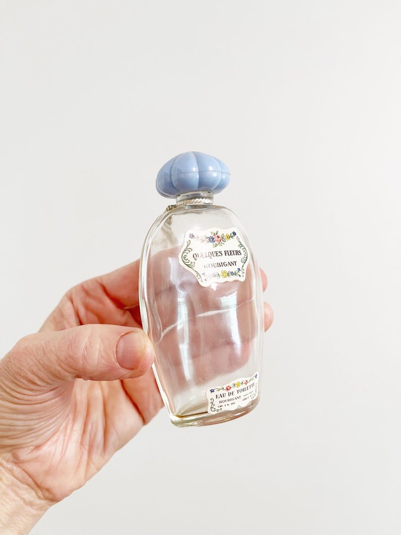 May include: A clear glass perfume bottle with a light blue cap. The bottle has a label with the words "Quelques Fleurs Houbigant" and "Eau de Toilette". The bottle is held in a hand.