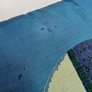 Pode incluir: Uma almofada retangular azul com uma superf&iacute;cie de tecido texturizado com pequenos fios puxados e desgaste vis&iacute;veis. No lado direito, &eacute; vis&iacute;vel parte de um design circular com um fundo verde claro e uma borda estampada em azul escuro e verde.