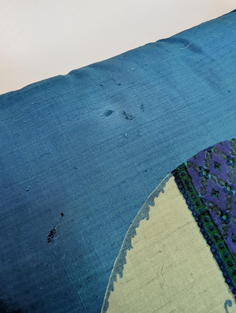 Pode incluir: Uma almofada retangular azul com uma superf&iacute;cie de tecido texturizado com pequenos fios puxados e desgaste vis&iacute;veis. No lado direito, &eacute; vis&iacute;vel parte de um design circular com um fundo verde claro e uma borda estampada em azul escuro e verde.