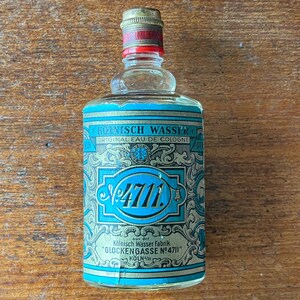 Puede incluir: Una peque&ntilde;a botella de vidrio transparente con una etiqueta azul y dorada. La etiqueta presenta el texto "4711" en una fuente grande azul y las palabras "K&ouml;lnisch Wasser Fabrik Glockengasse N&deg;4711" en una fuente dorada m&aacute;s peque&ntilde;a. La botella est&aacute; sobre una superficie de madera.