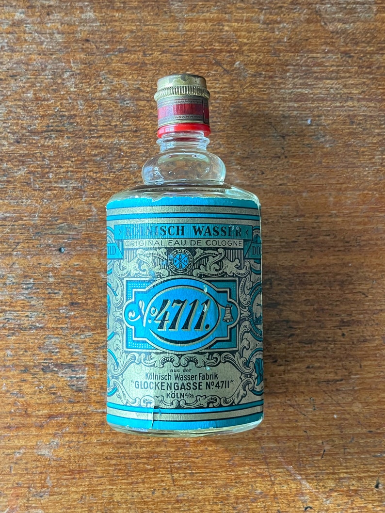 Puede incluir: Una peque&ntilde;a botella de vidrio transparente con una etiqueta azul y dorada. La etiqueta presenta el texto "4711" en una fuente grande azul y las palabras "K&ouml;lnisch Wasser Fabrik Glockengasse N&deg;4711" en una fuente dorada m&aacute;s peque&ntilde;a. La botella est&aacute; sobre una superficie de madera.