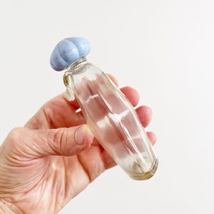 May include: A clear glass perfume bottle with a light blue, ribbed cap. The bottle has a slender, elongated shape and is held in a hand against a white background. A small charm hangs from the bottle's neck.
