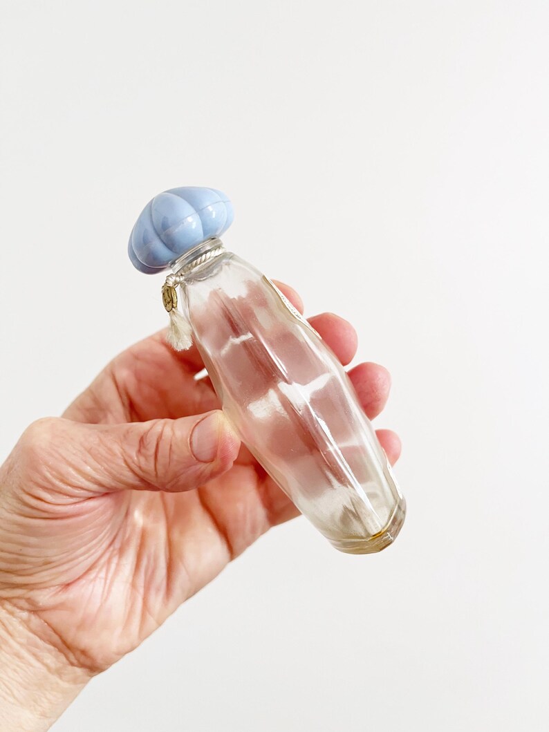 May include: A clear glass perfume bottle with a light blue, ribbed cap. The bottle has a slender, elongated shape and is held in a hand against a white background. A small charm hangs from the bottle's neck.