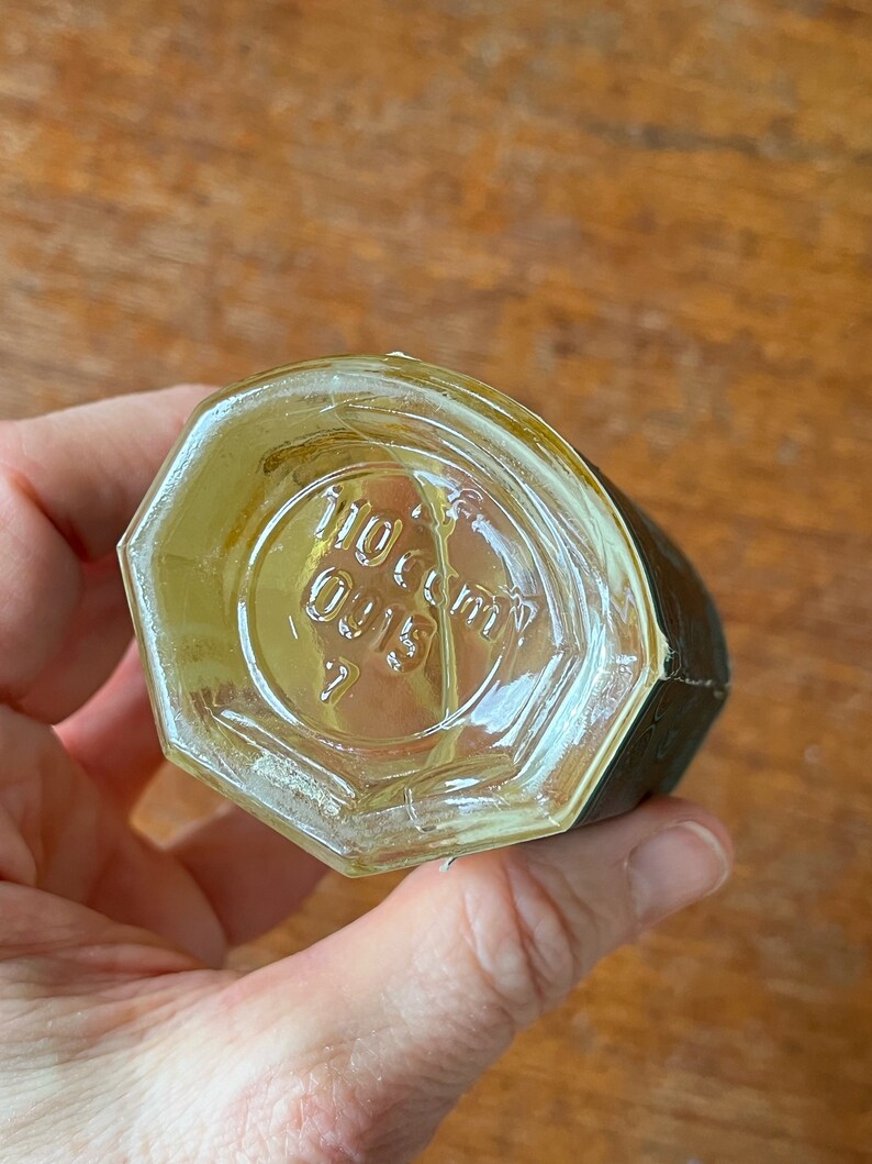 Puede incluir: Una botella de vidrio transparente con forma hexagonal y una etiqueta negra. La botella tiene una tapa de rosca y el texto "110 cc" y "Oil 15" es visible en la parte inferior de la botella.