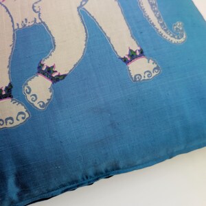 Pode incluir: Uma capa de almofada t&ecirc;xtil quadrada num tom azul vibrante com uma ilustra&ccedil;&atilde;o de um elefante branco. O elefante est&aacute; adornado com uma manta estampada em roxo e verde nas costas e bandas decorativas nos tornozelos, sobre um fundo azul s&oacute;lido.