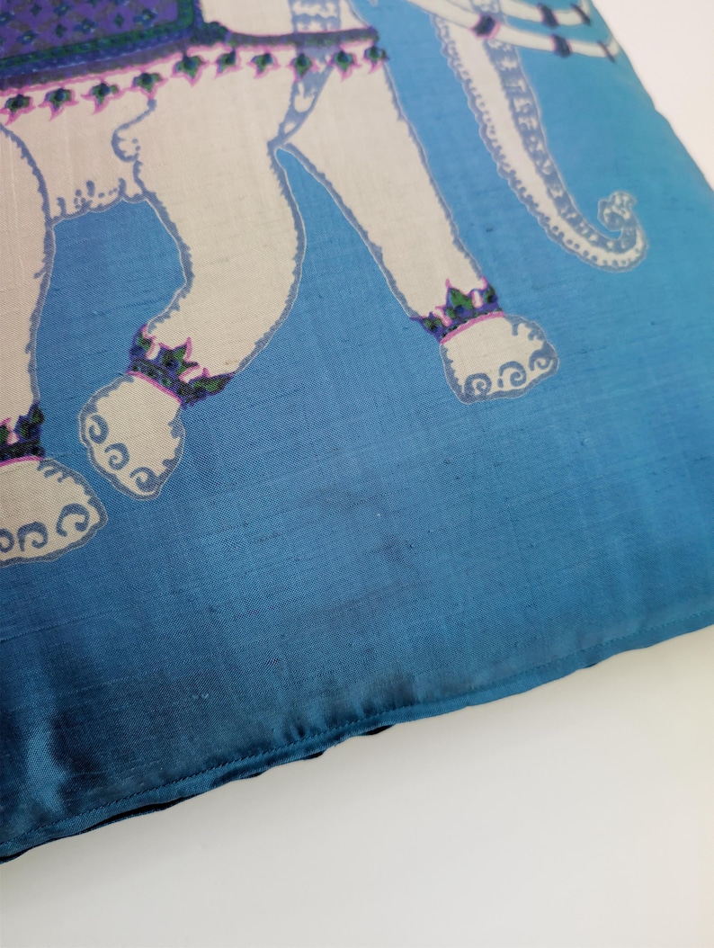 Pode incluir: Uma capa de almofada t&ecirc;xtil quadrada num tom azul vibrante com uma ilustra&ccedil;&atilde;o de um elefante branco. O elefante est&aacute; adornado com uma manta estampada em roxo e verde nas costas e bandas decorativas nos tornozelos, sobre um fundo azul s&oacute;lido.