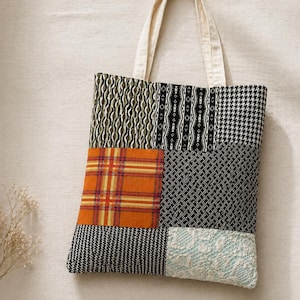 Può includere: Una borsa tote patchwork con manico color crema. La borsa presenta vari modelli di tessuto, tra cui pied-de-poule, scozzese e disegni geometrici in nero, bianco, arancione e verde. La borsa è realizzata in tessuto ed è progettata per trasportare oggetti.