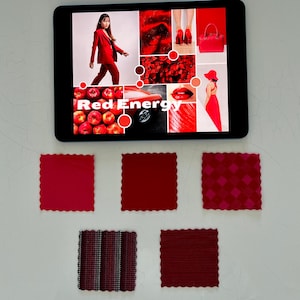 以下が含まれることがあります： タブレットには、衣類やアクセサリーなど、赤をテーマにした画像のコラージュが表示されています。タブレットの下には、さまざまなテクスチャと色合いの5つの赤い生地見本が白い表面に配置されています。タブレットの画面には「Red Energy」と表示されています。