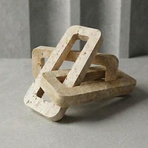 Puede incluir: Un objeto decorativo de eslabones de cadena tallado en piedra travertino beige. La pieza consta de tres eslabones rectangulares entrelazados con una superficie porosa y texturizada. Cada eslabón presenta esquinas redondeadas y un centro hueco, creando una escultura geométrica con variaciones de color natural moteado en tonos beige y crema en todo el material.