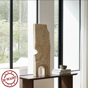 May include: A tall, rectangular beige travertine sculpture featuring carved, concentric semi-circular patterns and a hollow archway at the base. The stone has a porous, textured surface with natural variations in color and depth throughout the piece.
