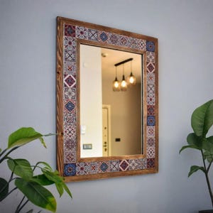 Puede incluir: Espejo rectangular con marco de madera y borde de azulejos decorativos. Los azulejos presentan un patrón repetitivo de diseños geométricos azules, rojos y blancos. El espejo refleja una habitación con luces colgantes y una puerta.