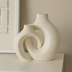 Könnte beinhalten: Zwei cremefarbene Keramikvasen mit gesprenkelter Textur. Die Vasen haben ein modernes, abstraktes Design, wobei eine Vase die andere teilweise umschließt. Die Vasen stehen auf einer weißen Oberfläche, mit einem strukturierten Kunstwerk im Hintergrund.