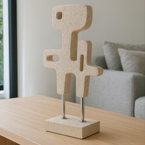 Könnte beinhalten: Abstrakte beige Skulptur mit einer geometrischen, menschenähnlichen Form. Die Skulptur hat einen rechteckigen Sockel, zwei Metallbeine und eine strukturierte Oberfläche. Sie steht auf einem hellbraunen Holztisch, im Hintergrund ein graues Sofa.