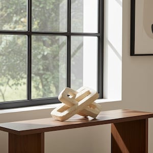 Puede incluir: Una escultura abstracta de color claro con un diseño tejido se encuentra sobre una mesa de madera marrón oscuro. La escultura está hecha de un material texturizado y está colocada frente a una ventana con marcos negros. El fondo incluye una vista de árboles.
