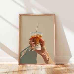 Op de afbeelding: Een ingelijste print van een ijskoffie in een doorzichtige plastic beker met een rietje. De koffie is gelaagd met een lichte basis en een donkerdere, karamelkleurige vloeistof erboven. De print zit in een houten lijst.