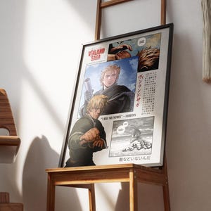 May include: Framed poster featuring artwork from "Vinland Saga" manga. The poster displays panels with characters and Japanese text. The color palette includes shades of brown, beige, and blue. The poster is propped on a wooden table.