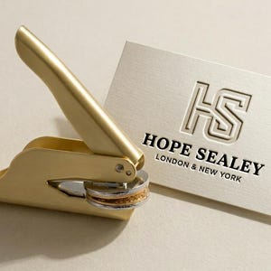 May include: A gold-colored seal press next to a cream-colored business card. The card features the embossed initials "HS" above the words "HOPE SEALEY LONDON & NEW YORK". The seal press is open, ready for use.