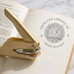 Op de afbeelding: Een goudkleurige lakstempel ligt op een open boek. De stempel staat naast een gedrukt ontwerp met de tekst "FROM THE LIBRARY OF HOPE SEALEY" met een illustratie van een boek, een veer en bladeren.