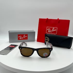 May include: Tortoiseshell-patterned sunglasses with brown lenses, displayed with a gray box, red shopping bag, black case, and cleaning cloth. The Ray-Ban logo is visible on the sunglasses, box, and bag.