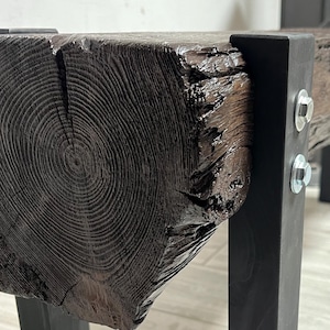May include: A dark stained wooden bench with black metal legs. The bench's wooden top shows visible grain. The bench is supported by four black metal legs. A fireplace is visible in the background. The bench is suitable for a living room.
