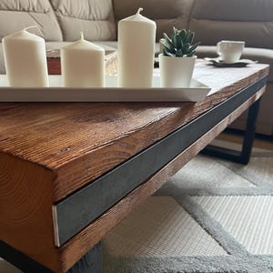 Könnte beinhalten: Rustikaler Couchtisch aus Holz mit dunklem Metallrahmen. Ein weißes Tablett enthält drei weiße Säulenkerzen und eine kleine Topf-Sukkulente. Der Tisch steht auf einem gemusterten Teppich.