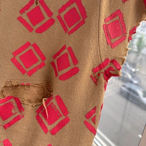 May include: Close-up of a tan-colored garment with a distressed design. The garment features a repeating pattern of bright red diamond shapes. The fabric has a worn, distressed look with visible tears and frayed edges.