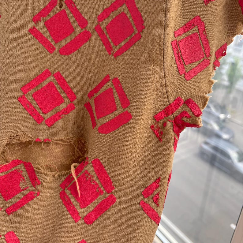 May include: Close-up of a tan-colored garment with a distressed design. The garment features a repeating pattern of bright red diamond shapes. The fabric has a worn, distressed look with visible tears and frayed edges.
