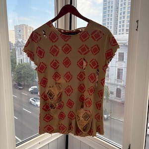 May include: Tan t-shirt with a red diamond pattern and distressed details. The short-sleeved shirt is hanging on a wooden hanger in front of a window with a city view. The shirt has several holes and frayed edges.
