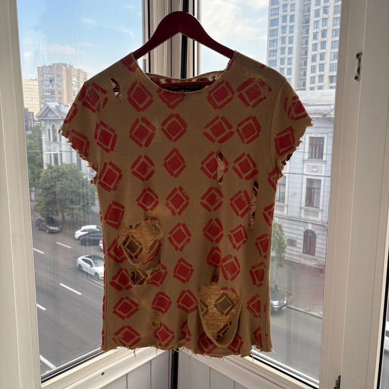 May include: Tan t-shirt with a red diamond pattern and distressed details. The short-sleeved shirt is hanging on a wooden hanger in front of a window with a city view. The shirt has several holes and frayed edges.