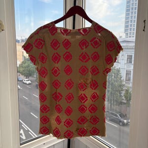 May include: Tan short-sleeved t-shirt with a repeating pattern of red diamond shapes. The shirt has a distressed look with several holes and frayed edges, hanging on a wooden hanger.