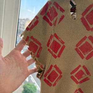 May include: Tan knit top with a red diamond pattern. The short sleeves have a distressed, frayed edge. The top is made of a textured knit fabric.