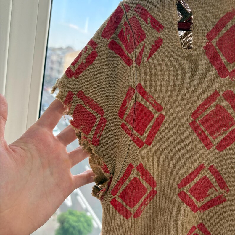 May include: Tan knit top with a red diamond pattern. The short sleeves have a distressed, frayed edge. The top is made of a textured knit fabric.