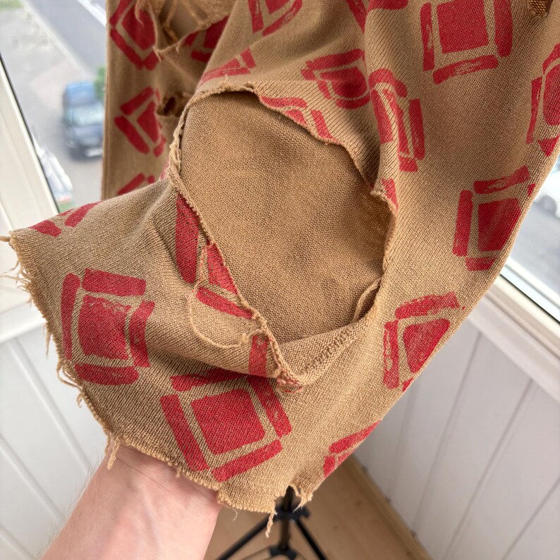 May include: Tan fabric with a repeating pattern of red squares. The fabric has distressed edges and visible tears, giving it a vintage or worn appearance. The fabric is likely part of a garment.