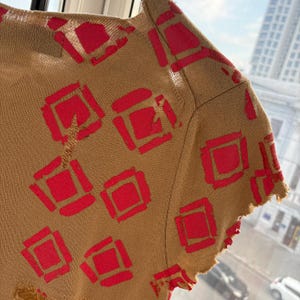 May include: Tan-colored knit top with a repeating pattern of bright red squares. The top has a distressed, frayed edge along the sleeve and hem. The fabric appears to be lightweight, possibly for warmer weather.