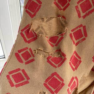 May include: Tan fabric with a repeating pattern of red diamond shapes. The fabric has several distressed holes, revealing a lighter tan material underneath. The design is visible on the fabric.