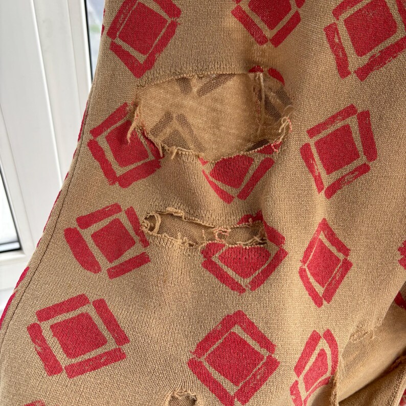 May include: Tan fabric with a repeating pattern of red diamond shapes. The fabric has several distressed holes, revealing a lighter tan material underneath. The design is visible on the fabric.