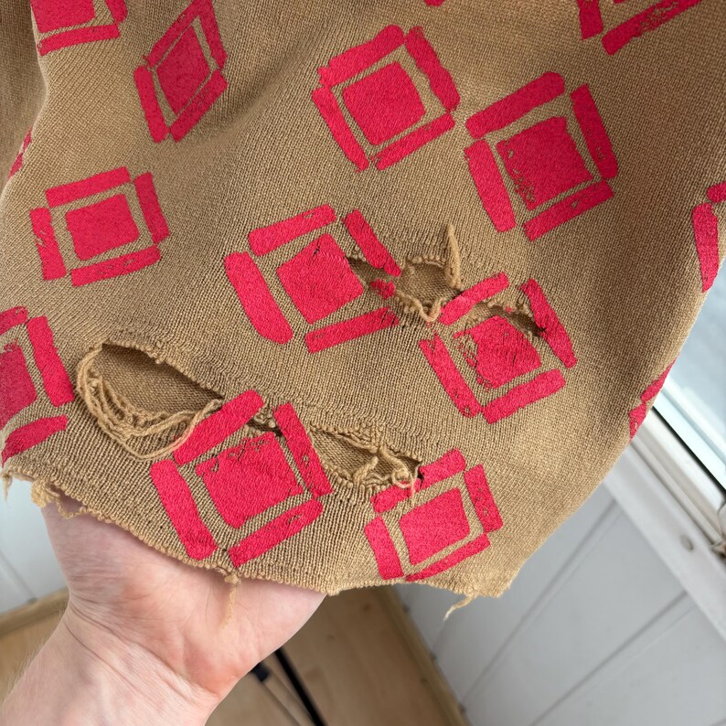 May include: Close-up of a tan fabric with a repeating pattern of red diamond shapes. The fabric has distressed holes along the edge, adding a vintage or worn aesthetic. The design is likely for apparel or textiles.