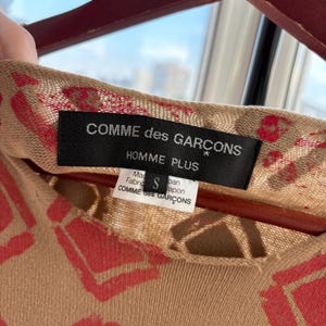May include: Close-up of a tan-colored garment with a red heart-shaped pattern. The black label reads "COMME des GARCONS HOMME PLUS" with a smaller tag below. The garment is size S and made in Japan.