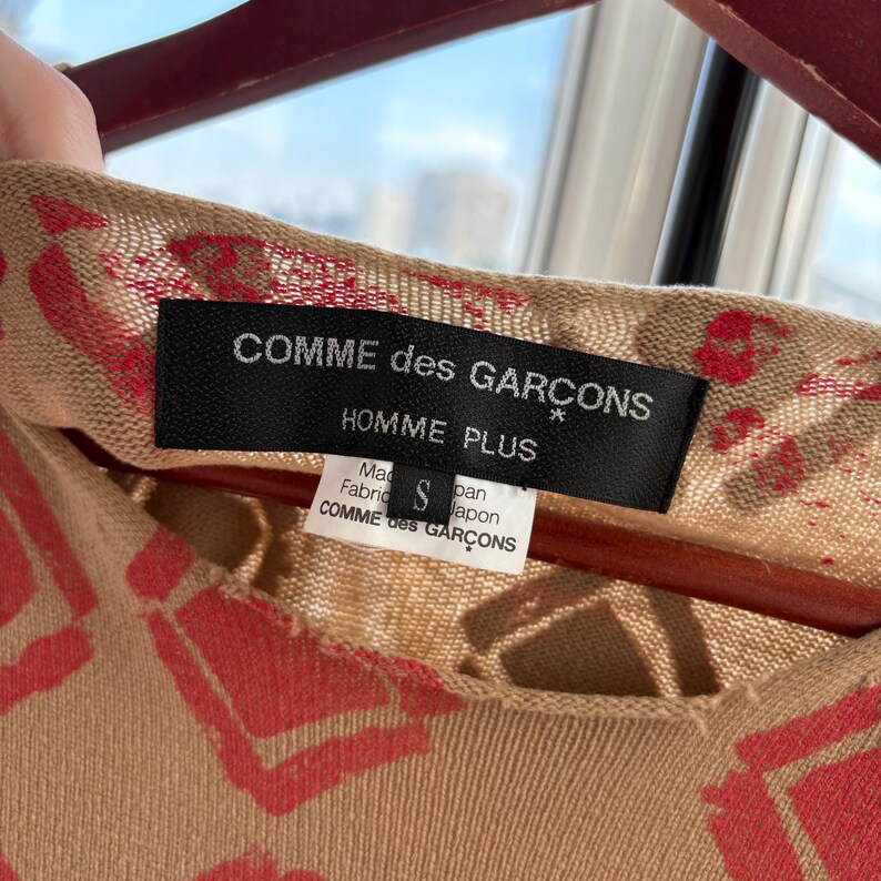 May include: Close-up of a tan-colored garment with a red heart-shaped pattern. The black label reads "COMME des GARCONS HOMME PLUS" with a smaller tag below. The garment is size S and made in Japan.