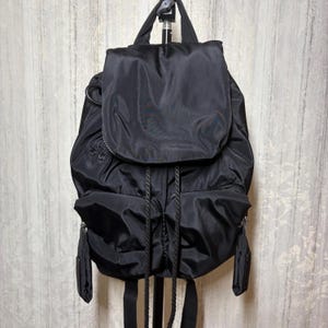 May include: A black nylon backpack featuring a large top flap, a drawstring closure with braided cords, and two front pockets with zip pulls. The bag includes a top handle and adjustable shoulder straps, providing a functional design.