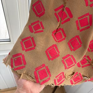 May include: Close-up of a tan fabric with a repeating pattern of red squares. The fabric has a distressed, frayed edge. The design is a bold, geometric pattern, creating a vintage or bohemian aesthetic.