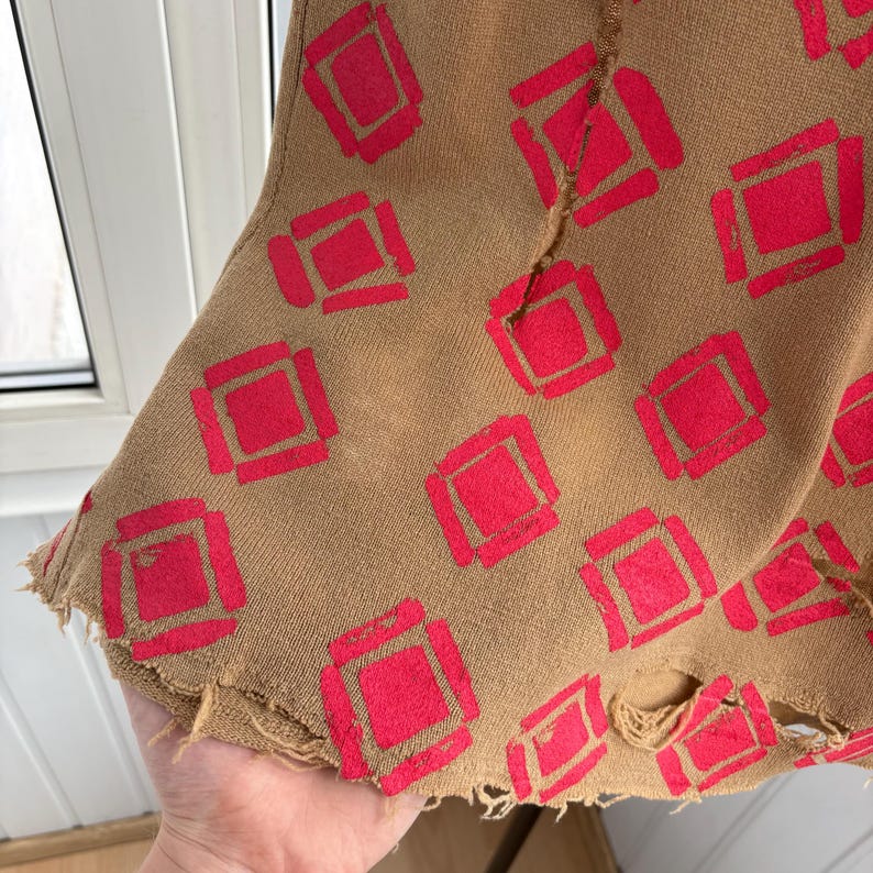 May include: Close-up of a tan fabric with a repeating pattern of red squares. The fabric has a distressed, frayed edge. The design is a bold, geometric pattern, creating a vintage or bohemian aesthetic.