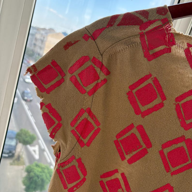 May include: Close-up of a tan-colored top with a repeating pattern of red squares. The top has a distressed, frayed edge on the sleeve. The fabric appears to be a knit or woven material. The design is a bold, geometric pattern.