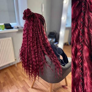 Peut inclure: Une personne avec de longs cheveux bouclés de couleur bordeaux, coiffés en chignon et en longues tresses. Les cheveux tombent dans le dos, avec un gros plan montrant une tresse détaillée avec des accents noirs. La personne est assise sur une chaise grise.