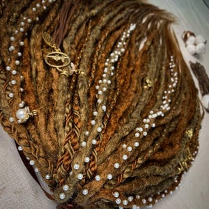 Puede incluir: Primer plano de rastas marrones y doradas adornadas con perlas blancas y detalles dorados. Las rastas están peinadas con una mezcla de texturas y colores, con una sección trenzada y dijes decorativos.