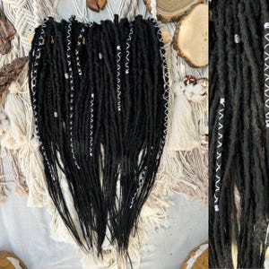 Könnte beinhalten: Schwarze Dreadlocks mit weißen Akzenten und dekorativen Perlen. Die Dreadlocks sind in einem Bündel angeordnet, wobei einige Strähnen locker hängen. Der Stil zeichnet sich durch aufwendige weiße Nähte und kleine silberne Perlen aus.