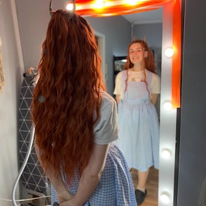 Puede incluir: Una persona con largo cabello rojizo ondulado y un accesorio para el cabello en forma de cuerno se encuentra frente a un espejo iluminado. La persona lleva una camiseta blanca y un vestido de cuadros azul y blanco. El espejo refleja el rostro sonriente.