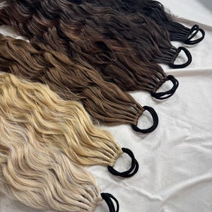 Könnte beinhalten: Vier Bündel gewellter Haarverlängerungen in verschiedenen Braun- und Blondtönen, jeweils mit einem schwarzen Gummiband befestigt. Die Farben reichen von Dunkelbraun bis Hellblond, mit einer Mischung aus Tönen.
