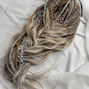 Op de afbeelding: Een set blonde, bruine en grijze synthetische dreadlocks, gestyled met vlechten en losse strengen. De dreadlocks zijn versierd met zilveren halve maan bedels en strengen kleine witte kralen. Een zwart lint zet het geheel vast.