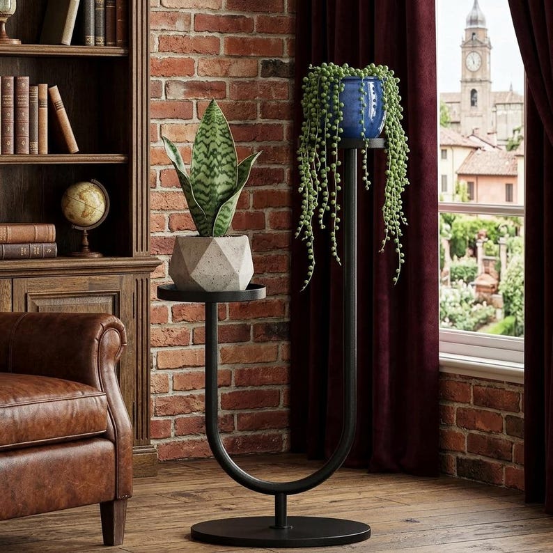 Op de afbeelding: Een U-vormige plantenstandaard van zwart metaal met twee ronde niveaus. Het onderste niveau bevat een geometrische betonnen pot met een sansevieria, terwijl het bovenste niveau een blauwe keramische pot met een erwtenplant ondersteunt.
